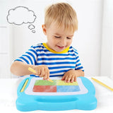 Un enfant de 5 ans qui est assis sur une table blanche et qui dessine sur une ardoise magique bleu 4 carrés bleu, vert, jaune et rouge avec un stylet. Il porte un tee-shirt à rayure bleu et blanc. Il a une bulle vide au dessus de la tête